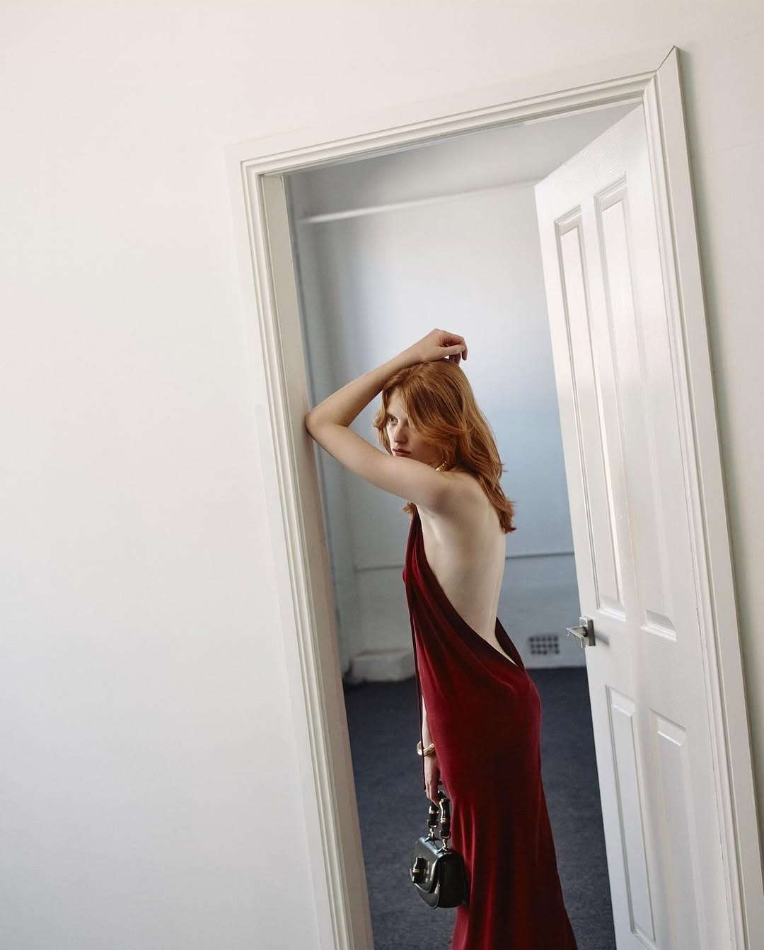 An adult female with red hair wearing a red open back gown and holding a handbag leans against a white doorway in an empty room.