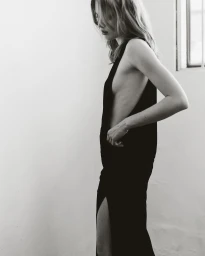 Model in a black side-slit dress standing in profile against a plain white interior wall