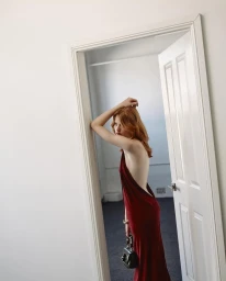 Red-haired model in a long burgundy backless dress holding a dark handbag in a doorway.