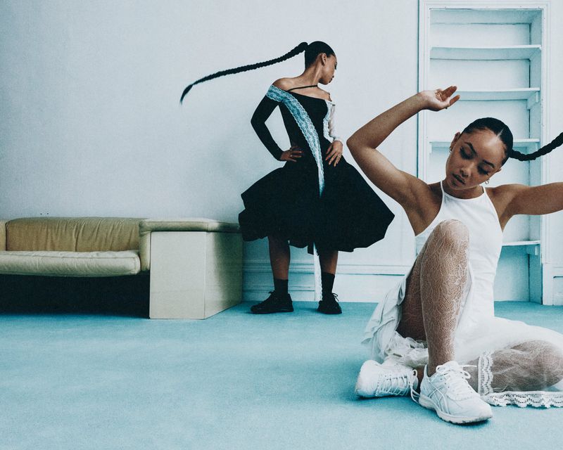 Two women in fashionable dresses and sneakers pose in a light blue indoor setting with minimal furniture and a clean, contemporary aesthetic.