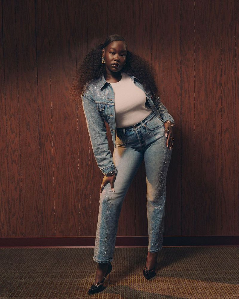 A striking portrait of an African American woman in a denim jacket and jeans, confidently posing against a wood-panelled wall.