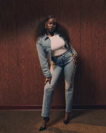 A striking portrait of an African American woman in a denim jacket and jeans, confidently posing against a wood-panelled wall.