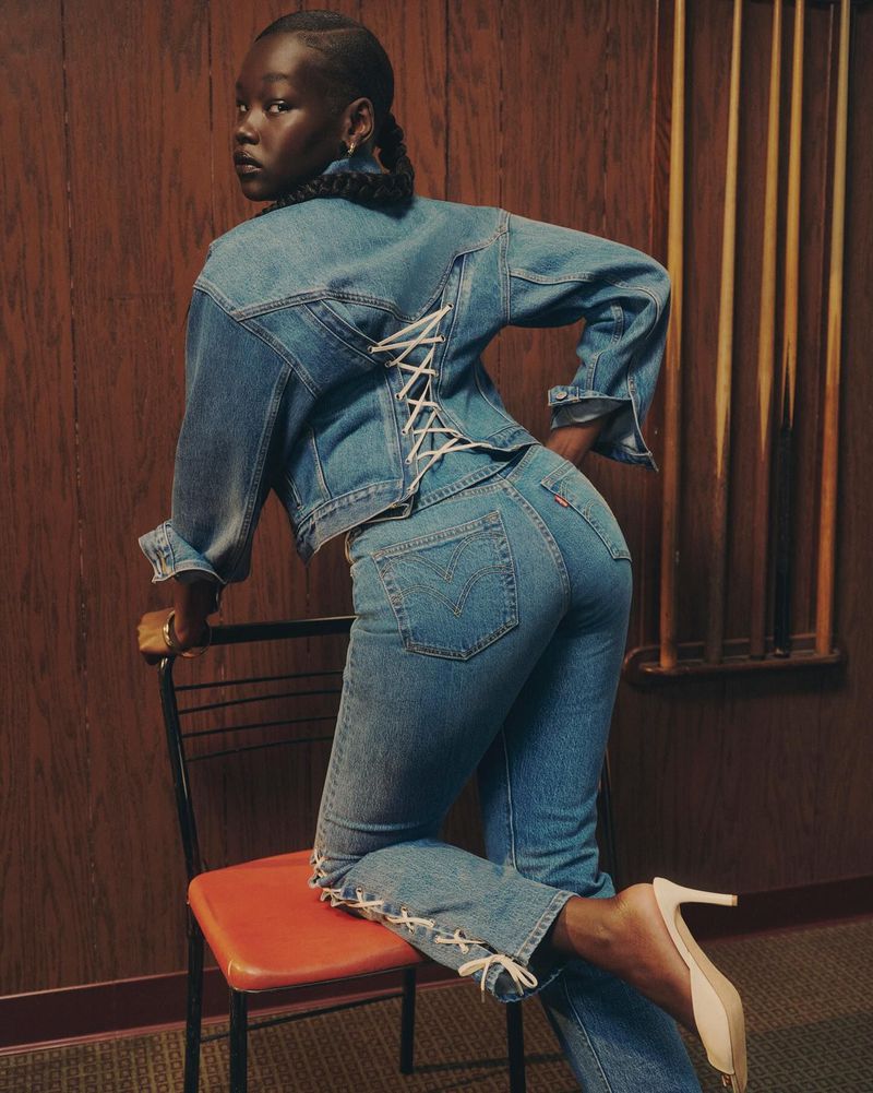 Woman poses in denim jacket and jeans with laced details, standing next to a chair with a wood panel background, fashion shoot.