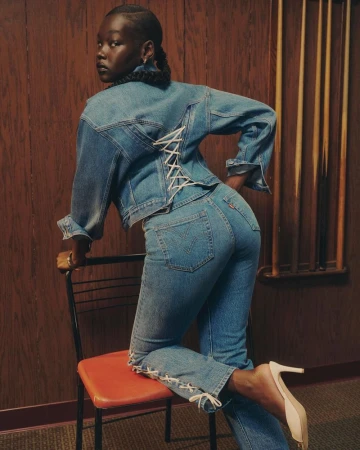 Woman poses in denim jacket and jeans with laced details, standing next to a chair with a wood panel background, fashion shoot.