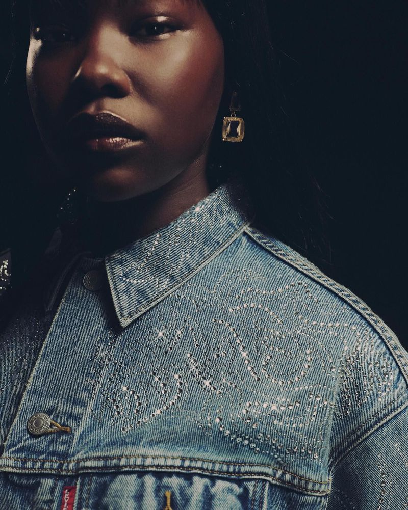 A dark-skinned woman wearing an embellished denim jacket and gold earrings poses in front of a dark background in a studio shot.