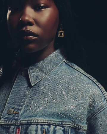 A dark-skinned woman wearing an embellished denim jacket and gold earrings poses in front of a dark background in a studio shot.