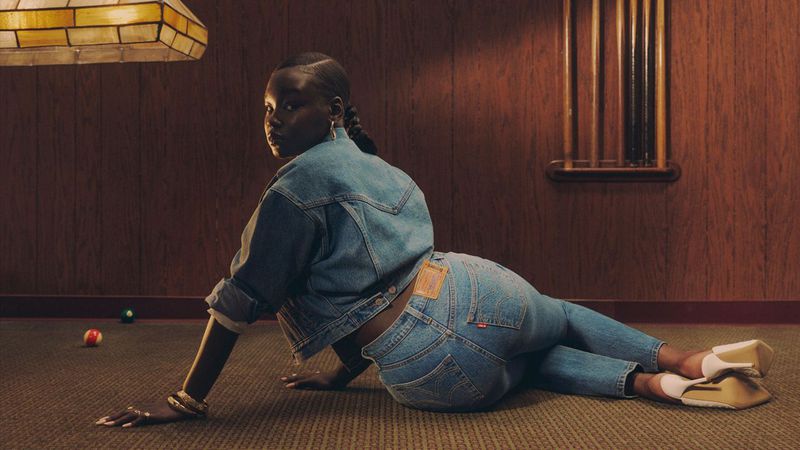A Black woman with braided hair poses on the floor wearing a denim jacket and jeans, accessorized with rings and bracelets in a warm indoor setting.