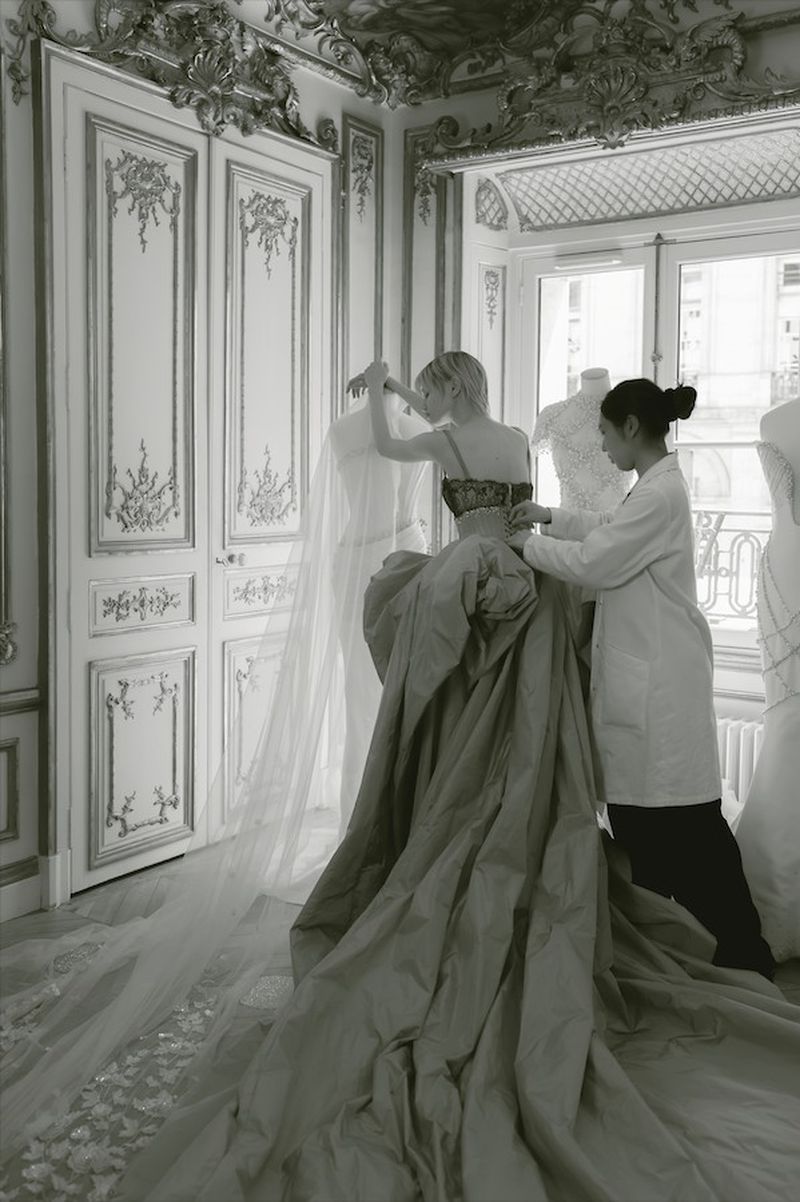 Two dressmakers fitting a bespoke couture gown on a mannequin in an ornate studio, showcasing detailed design & craftsmanship.