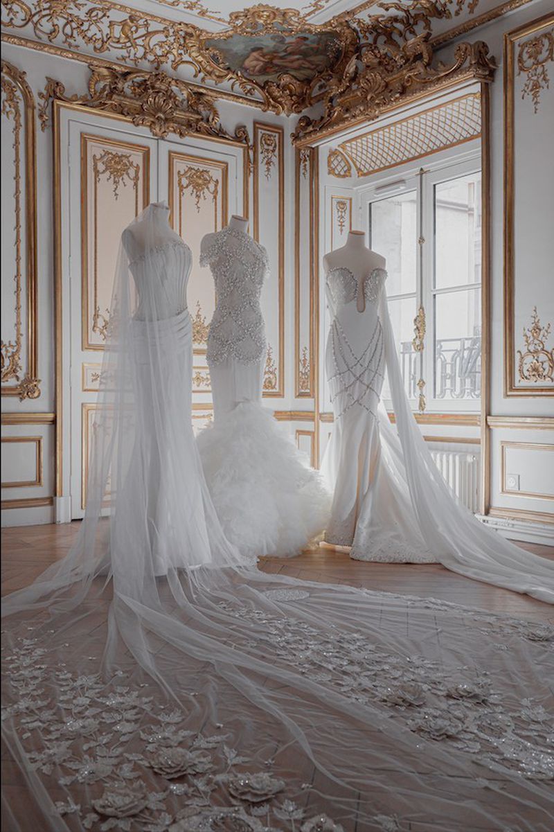 Three wedding dresses on mannequins are showcased in an ornate room with gold trim and large windows, highlighting bridal fashion and interior elegance.