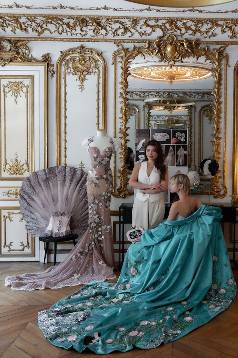 Two women in a fashion studio with ornate gilded decor, surrounded by couture gowns. One woman is seated, draped in a blue dress.