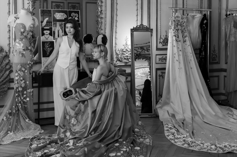 Two women in a vintage interior with ornate gilded details, wearing elaborate dresses with floral embroidery and tulle accents.