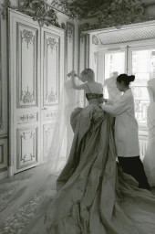 An artisan in a white coat adjusts a voluminous gown on a model in an ornate showroom with tall panelled doors and large windows.
