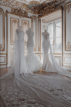 Three white bridal couture gowns on mannequins in an ornate gilded room with an embroidered veil trailing on the parquet floor.