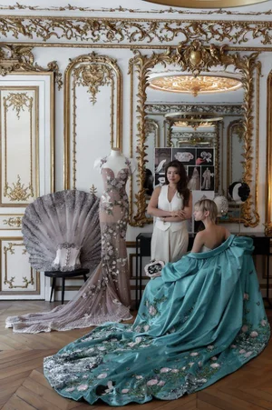 Two women posing in an ornate room with a mannequin displaying an embroidered gown and a model in a voluminous teal dress