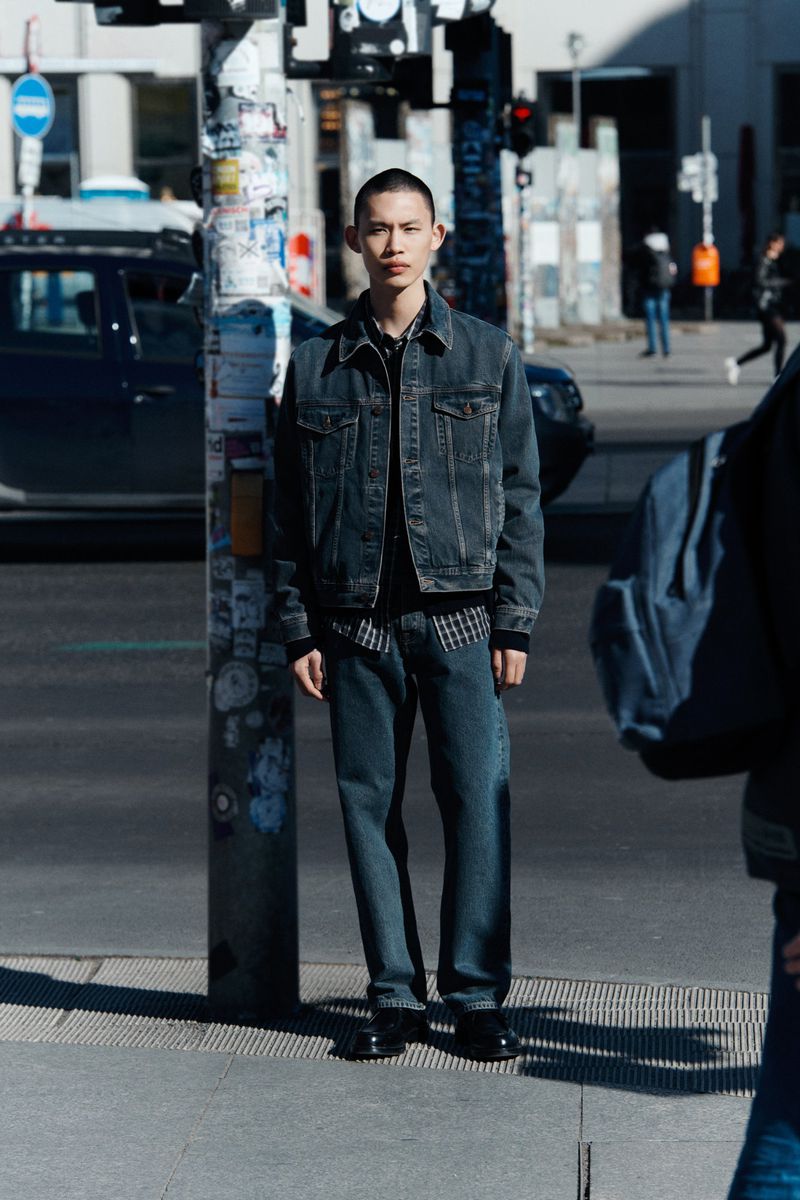 Young man in denim jacket and jeans stands on city street by a pole; dark denim outfit, urban background, daytime lighting, fashion pose.