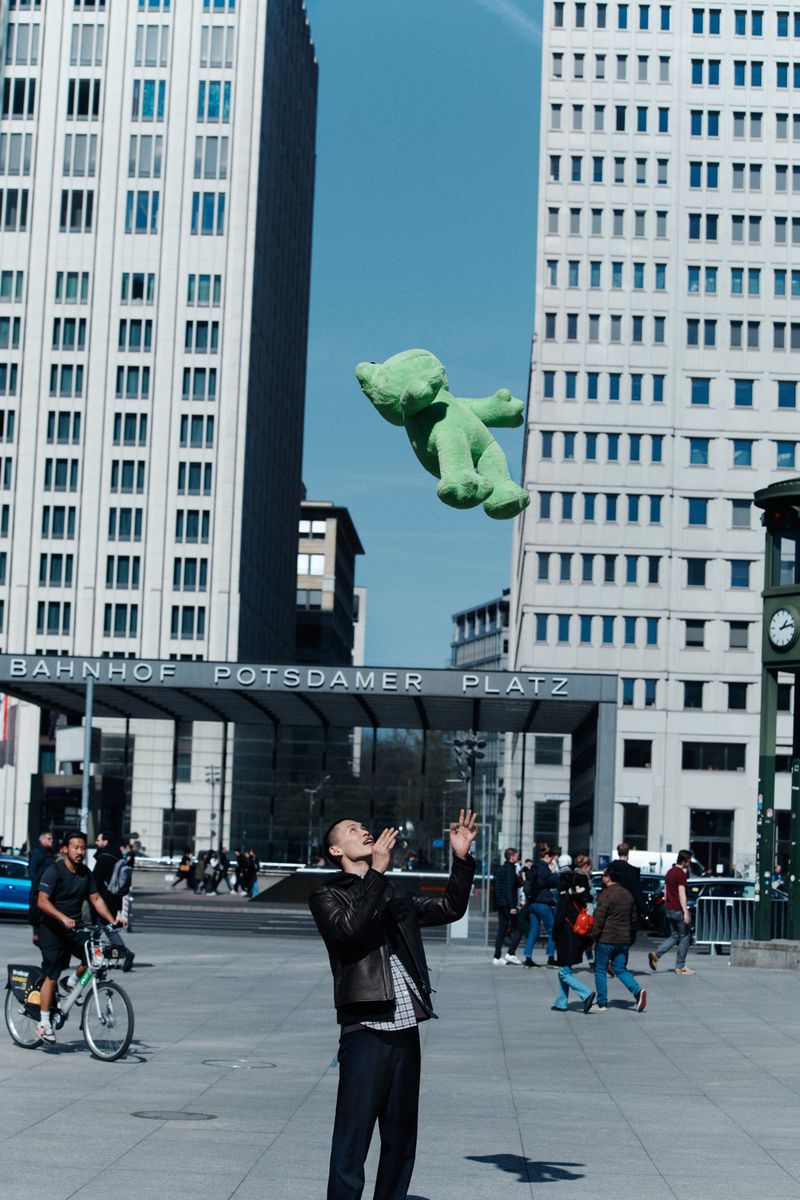 Man wearing a black leather jacket stands in an urban square, tossing a green plush bear in the air near train station and modern buildings.