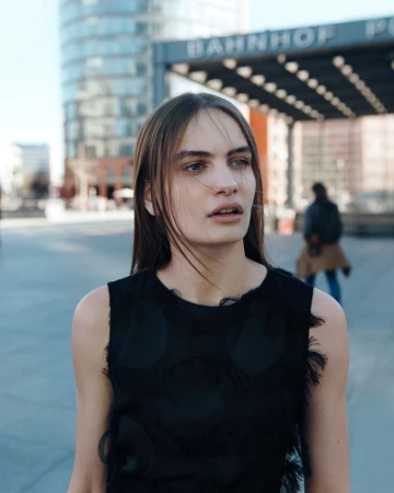 A young woman stands in an urban setting with modern buildings, captured in a medium shot with a shallow depth of field.