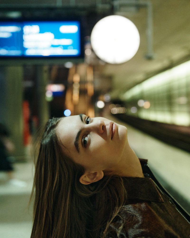 Young woman poses indoors in a medium shot, looking upward with a blurred background featuring architectural elements and lighting fixtures.