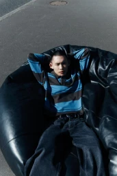 Model relaxing on a black leather beanbag wearing a blue striped polo shirt and dark jeans against an asphalt background.