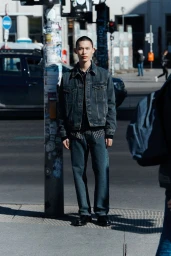 Model standing on a city street next to a pole covered in stickers, wearing a denim jacket and jeans.