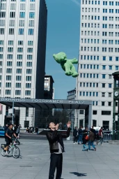 Man in a black leather jacket throws a large green teddy bear into the air at Potsdamer Platz under a clear blue sky.