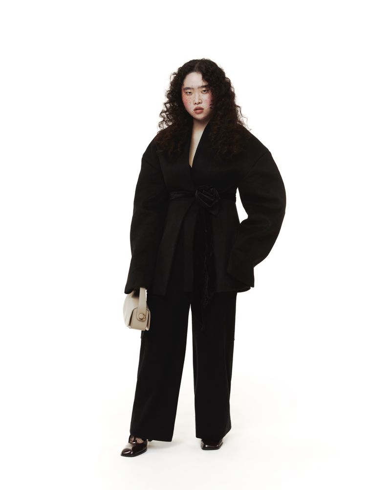 Full-length shot of an adult woman posing in a black belted jacket and matching pants, set against a stark white backdrop in a photo studio.
