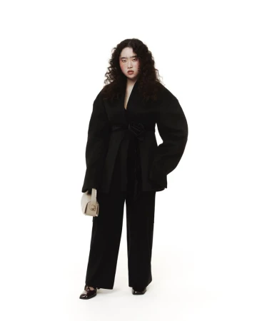 Full-length shot of an adult woman posing in a black belted jacket and matching pants, set against a stark white backdrop in a photo studio.
