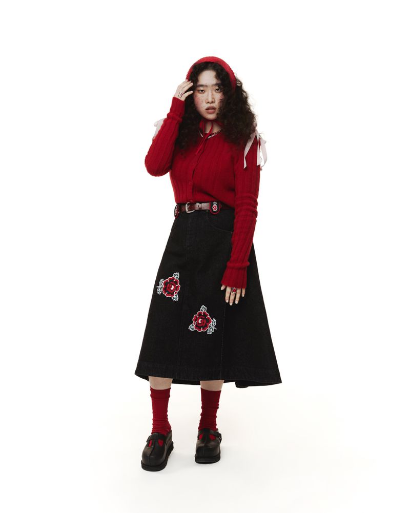 A stylish woman in a red sweater and black skirt with floral embroidery poses confidently against a bright white backdrop in a studio setting.