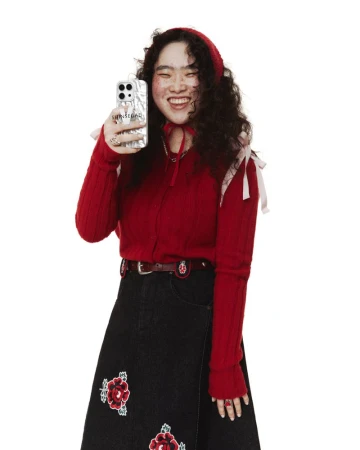 A young woman poses in a red knit sweater, black skirt with floral embroidery, and matching red knit cap while holding a phone.