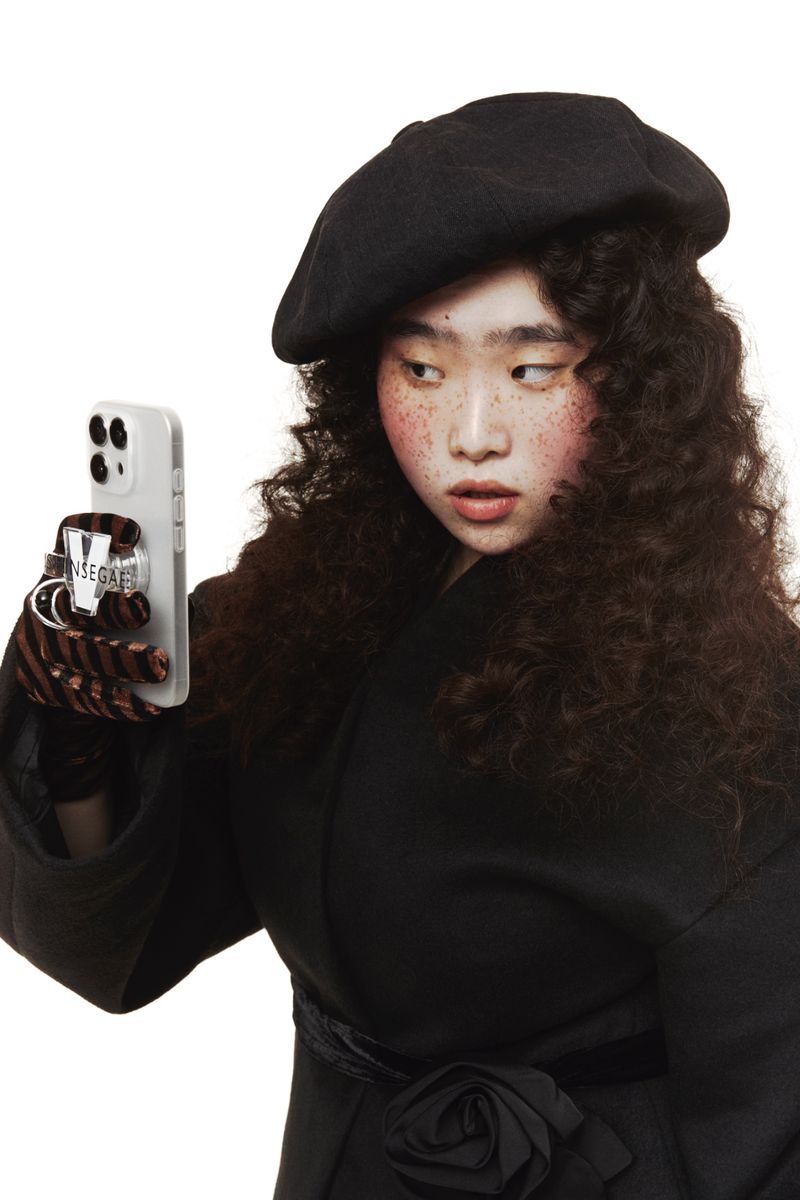 Young woman with curly hair and beret poses indoors holding a smartphone with a clear case in front of a stark white background.