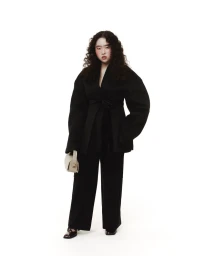 Model Yoorim Bae wearing an oversized black belted jacket and wide-leg trousers while holding a beige handbag against a white background.