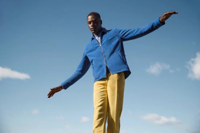 A Black man balances with arms outstretched against a blue sky, wearing a blue jacket and yellow pants, captured in natural daylight.