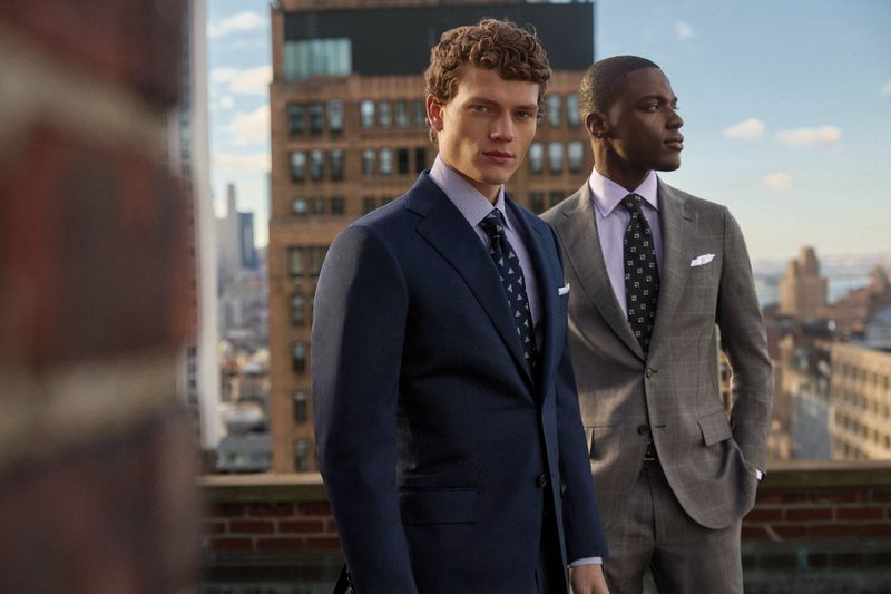 Two men stand on a rooftop wearing formal suits, showcasing fashion against a cityscape backdrop, captured in medium shot with a clear sky.