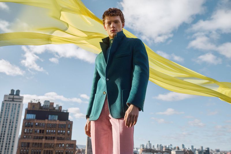 A fashion shot featuring a male model wearing a green blazer and pink pants, posing against a blue sky with a yellow fabric overlay.