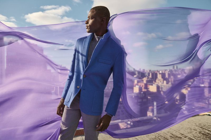 An adult man in a blazer and slacks poses against a city skyline, with a sheer violet fabric floating around him in an outdoor setting.