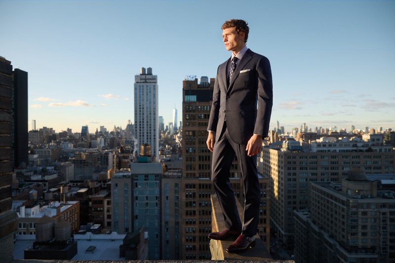Adult male fashion model poses in a suit on a rooftop overlooking a city skyline, featuring modern architecture against a clear sky.