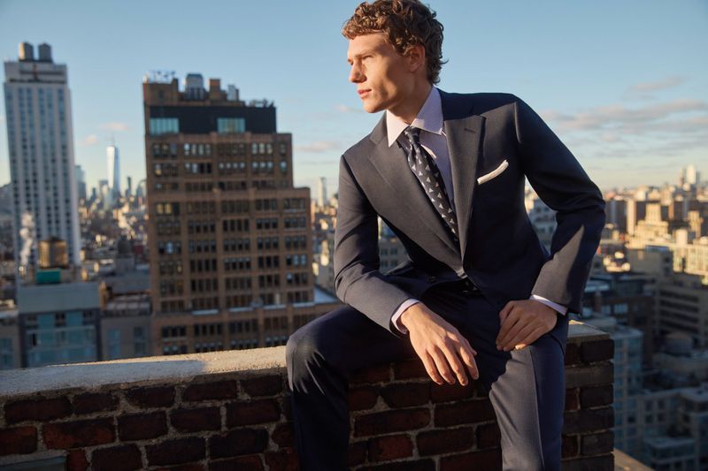A man in a navy suit sits atop a brick ledge with a city skyline backdrop, bathed in soft light, showcasing a blend of style and urban scenery.
