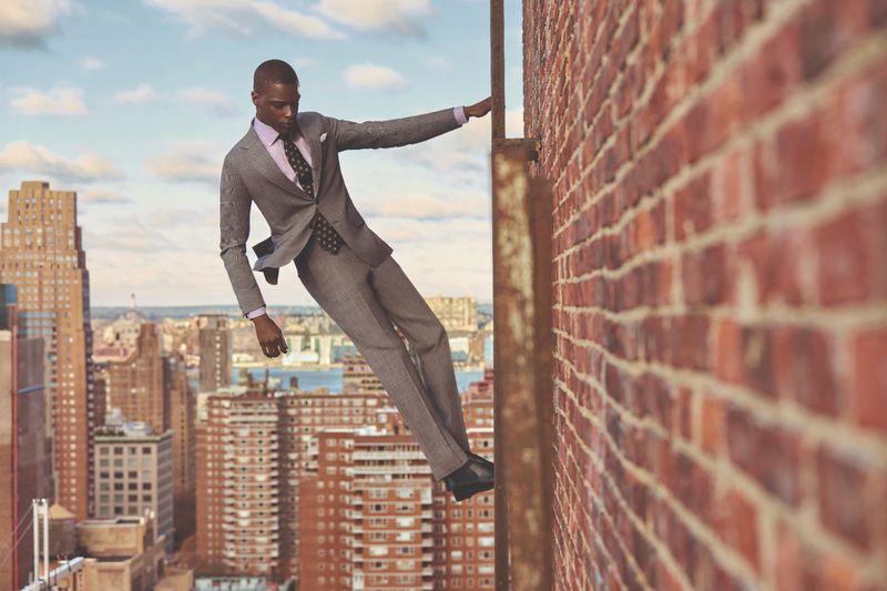 A stylish Black man in a grey suit poses on a red brick rooftop, with a city skyline backdrop and blue sky with clouds.