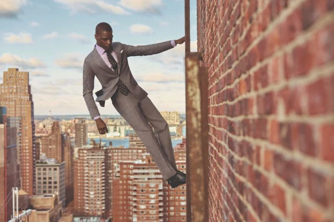 A stylish Black man in a grey suit poses on a red brick rooftop, with a city skyline backdrop and blue sky with clouds.