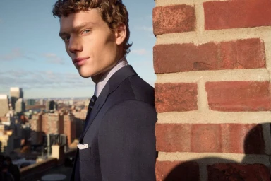 Close-up portrait of a male model in a navy suit leaning against a brick column with a city skyline backdrop.