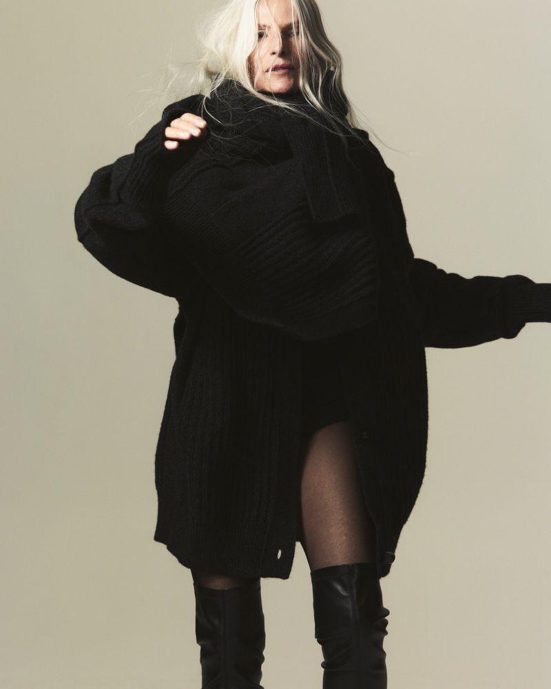 A mature adult woman with gray hair is posing in a flowing black sweater dress and black boots against a neutral studio backdrop.