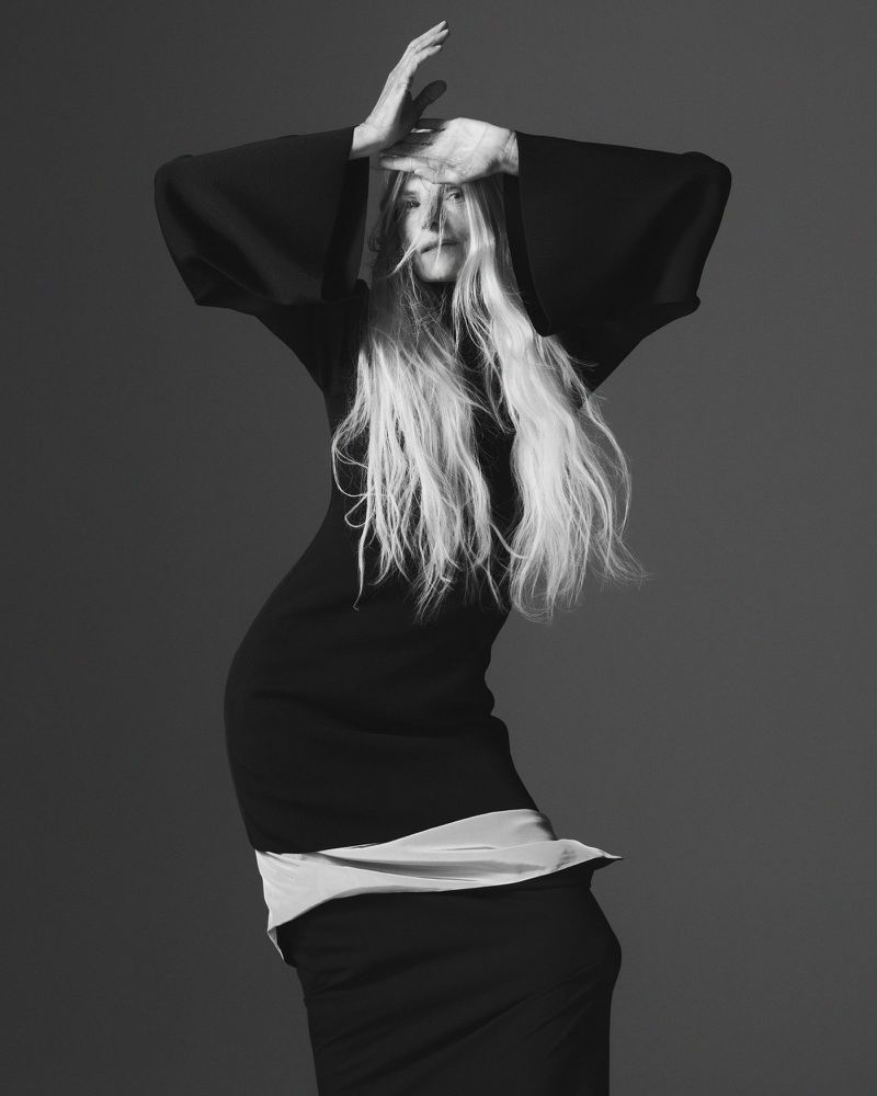 Monochrome portrait of a blonde woman with hands raised over her head, wearing a dark top and skirt against a gray backdrop in a studio.