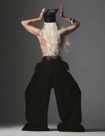 Back view of adult with long white hair and black hat, wearing loose black pants and a silver bracelet in a grey studio setting.
