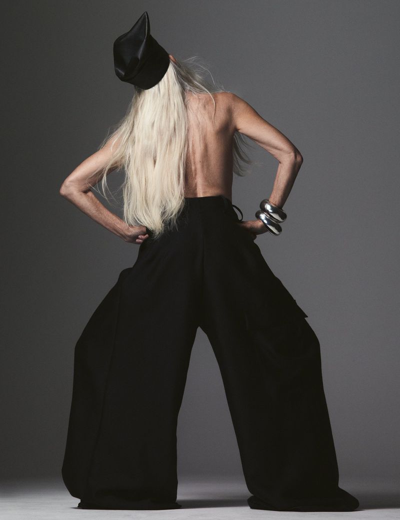 Back view of a slender, mature adult female posing in a photo studio wearing a dark-colored top and wide leg pants with stylish bracelet.