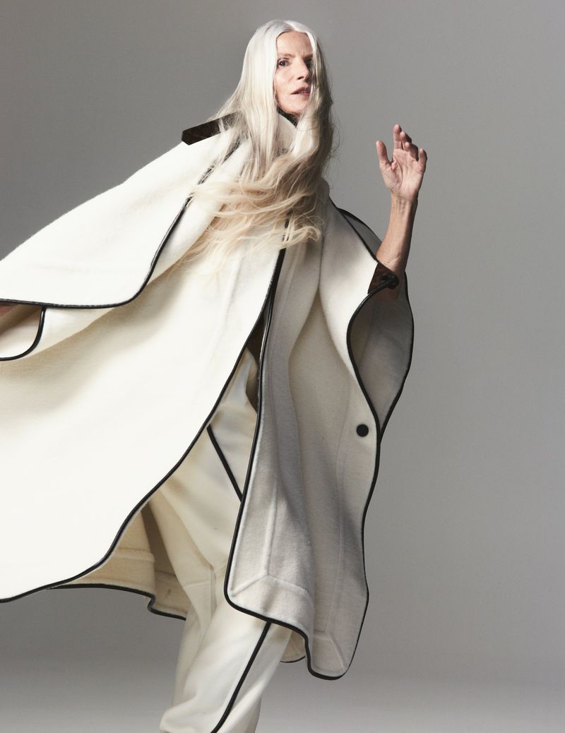 Elegant older woman in a white cape and matching pants with black trim strikes a pose in a studio. Fashion shot on neutral backdrop.