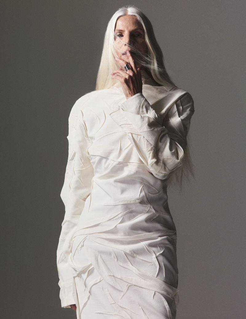 Woman with long white hair in a white dress poses in a studio with a finger to her lips, creating a serene and fashionable portrait.