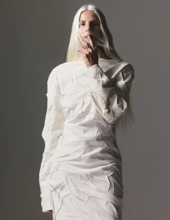 Woman with long white hair in a white dress poses in a studio with a finger to her lips, creating a serene and fashionable portrait.