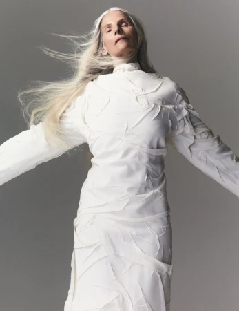 An older adult female with long gray hair and closed eyes poses indoors wearing a textured white dress against a grey background.