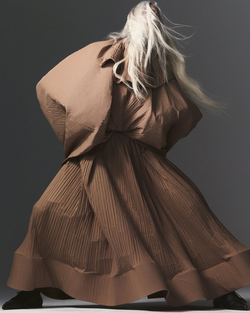 Fashion shot featuring an adult female model in a dramatic pose wearing a large, textured, brown, pleated dress with long blonde hair.