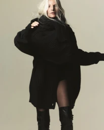 Model in black turtleneck sweater and tall boots posing against a neutral grey background
